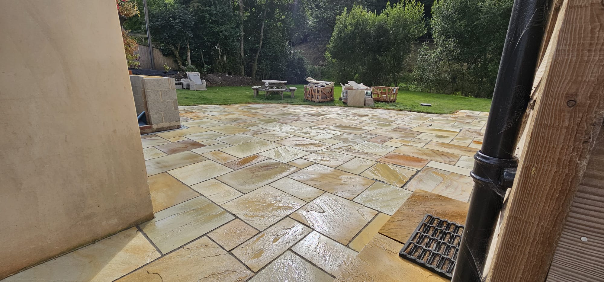 Wet Indian sandstone paving between a rendered wall and a timber-clad building, with a drainage grate