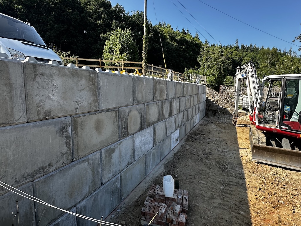 A long interlocking concrete block retaining wall under construction with an excavator alongside
